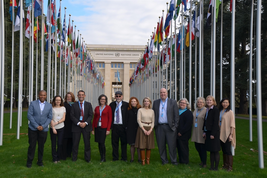 [ai] A group of 12 individuals standing in front of the United Nations building in Geneva, surrounded by flagpoles displaying various national flags. The participants are dressed in professional attire.
